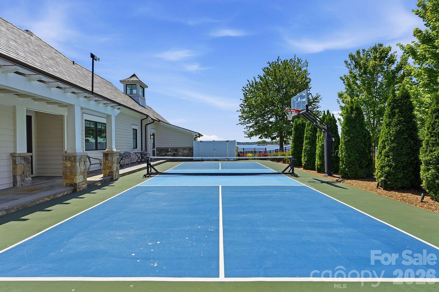 This outdoor space features a vibrant blue pickleball court situated directly adjacent to the side of a light-colored home with stone accents. A basketball hoop stands at the edge of the court, which is bordered by a row of tall, manicured evergreen trees and offers a scenic view of a lake in the distance. The perspective is a wide-angle shot from the edge of the court, emphasizing the recreational amenities and the peaceful waterfront setting.