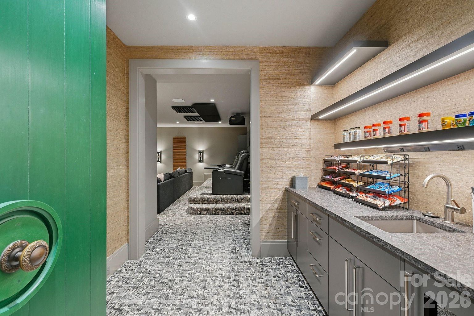 This image captures a wet bar area leading into a home theater, showcasing a sophisticated entertainment space. The bar features dark cabinetry, a stone countertop with an integrated sink, and open shelving stocked with snacks and drinks, while the adjacent theater room is visible through a wide doorway, revealing tiered seating and a dark, patterned carpet. The perspective is from the threshold of the bar area, looking toward the theater, highlighting the seamless transition between the snack station and the viewing area.