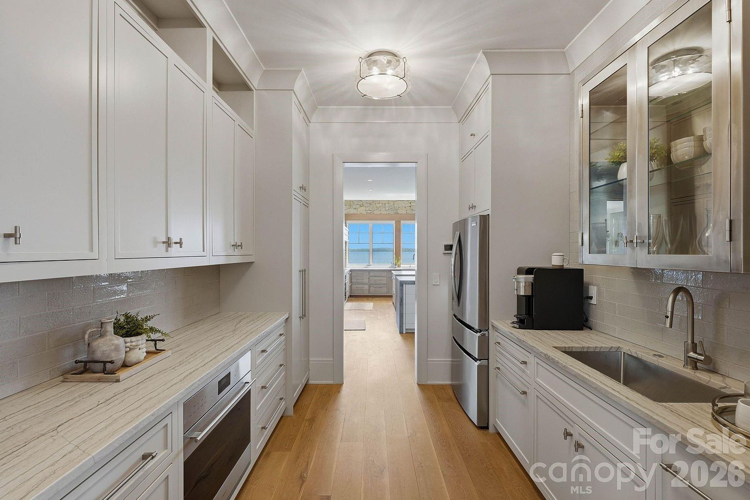 This elegant galley-style kitchen features crisp white cabinetry, light-toned stone countertops, and a sleek stainless steel sink and appliances. The space is illuminated by a central flush-mount light fixture and leads directly into an adjacent room with a view of the water through large windows. The light wood flooring and neutral color palette create a bright, sophisticated, and airy atmosphere.