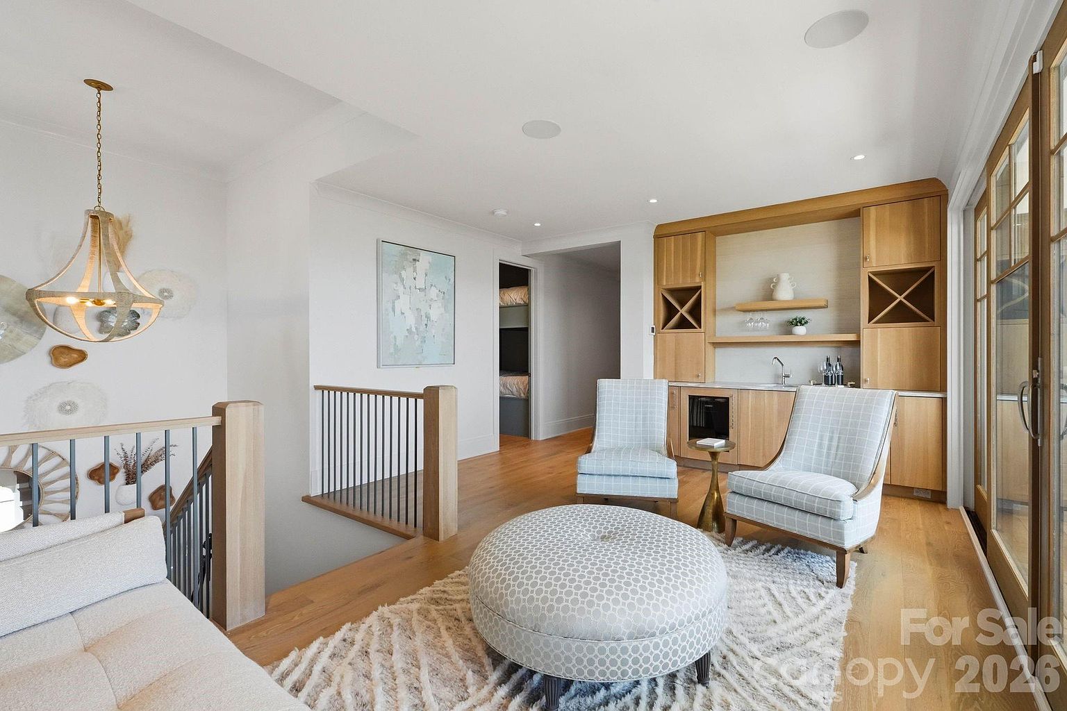 This inviting loft-style living area features light wood flooring, a textured area rug, and a central circular ottoman. A built-in light wood wet bar with shelving and cabinetry provides a sophisticated focal point, complemented by two plaid armchairs. The space is open to a staircase with a metal railing and offers a glimpse into an adjacent room, creating a bright and airy atmosphere.