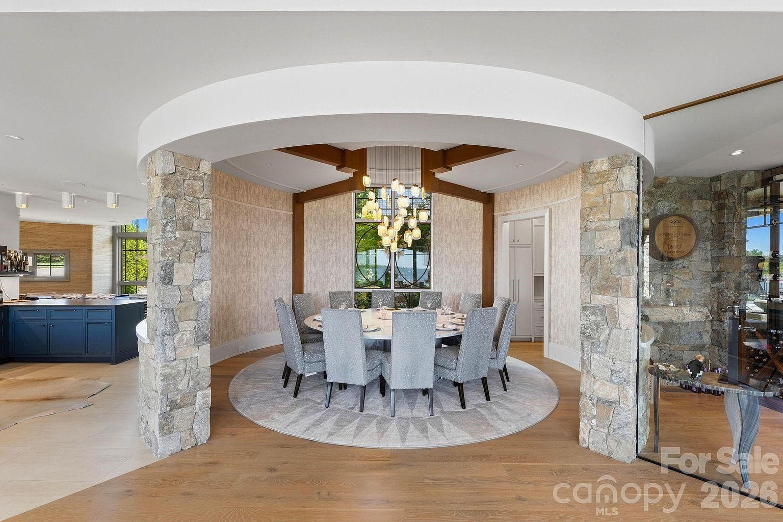 This elegant dining room features a circular layout defined by two prominent stone-clad pillars and a curved architectural ceiling detail. A large round table with eight upholstered chairs sits beneath a striking multi-pendant chandelier, while a circular area rug anchors the space on light wood flooring. The room is framed by a large window looking out to greenery, creating a sophisticated and inviting atmosphere for formal gatherings.