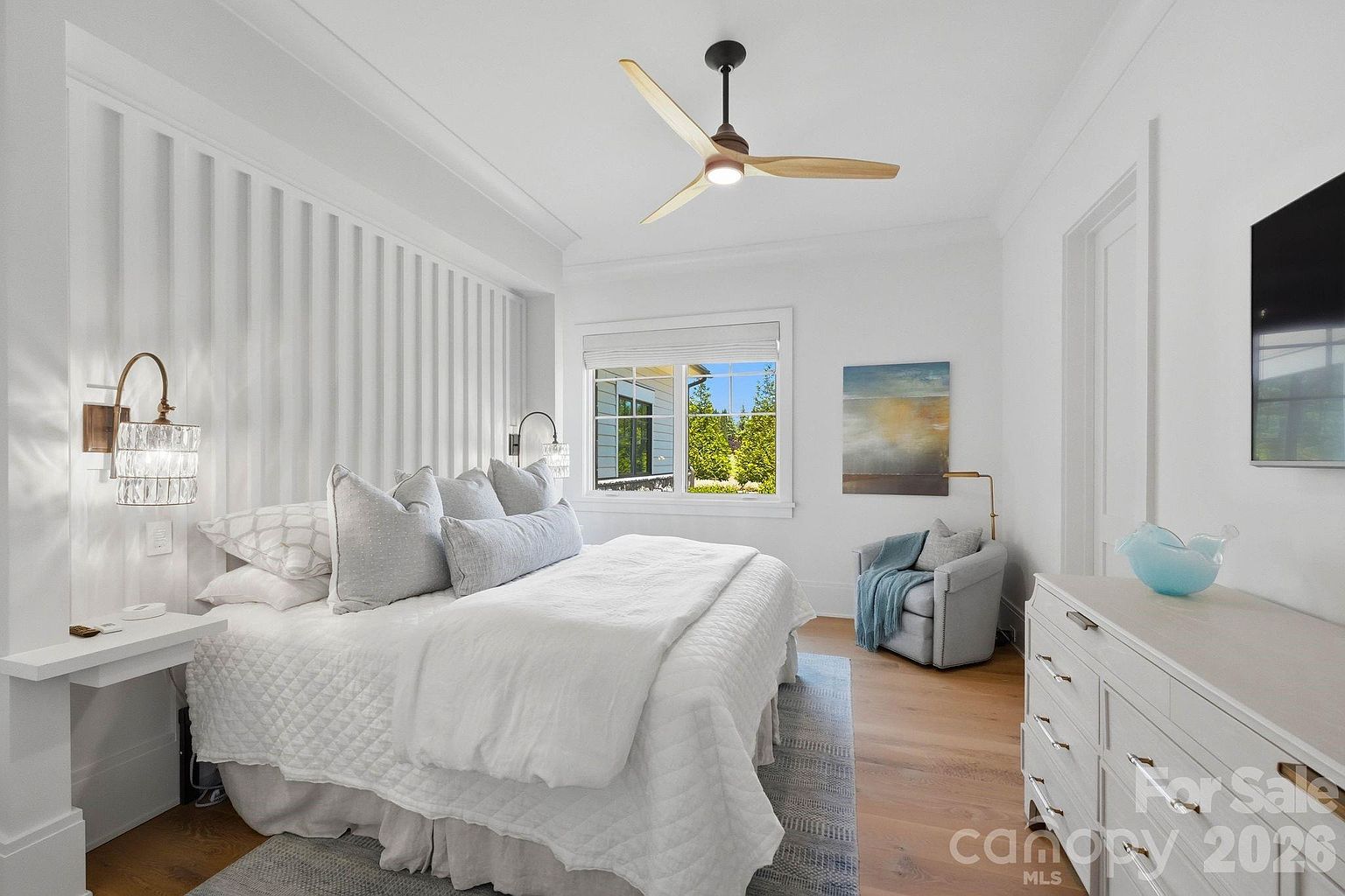 This bright and airy primary bedroom features a textured, vertical-paneled accent wall behind a plush bed dressed in white linens. The room is anchored by light wood flooring and a patterned area rug, with a cozy armchair tucked into the corner near a window that provides natural light. A white dresser with metallic hardware and a wall-mounted television complete the space, creating a clean, modern, and inviting atmosphere.