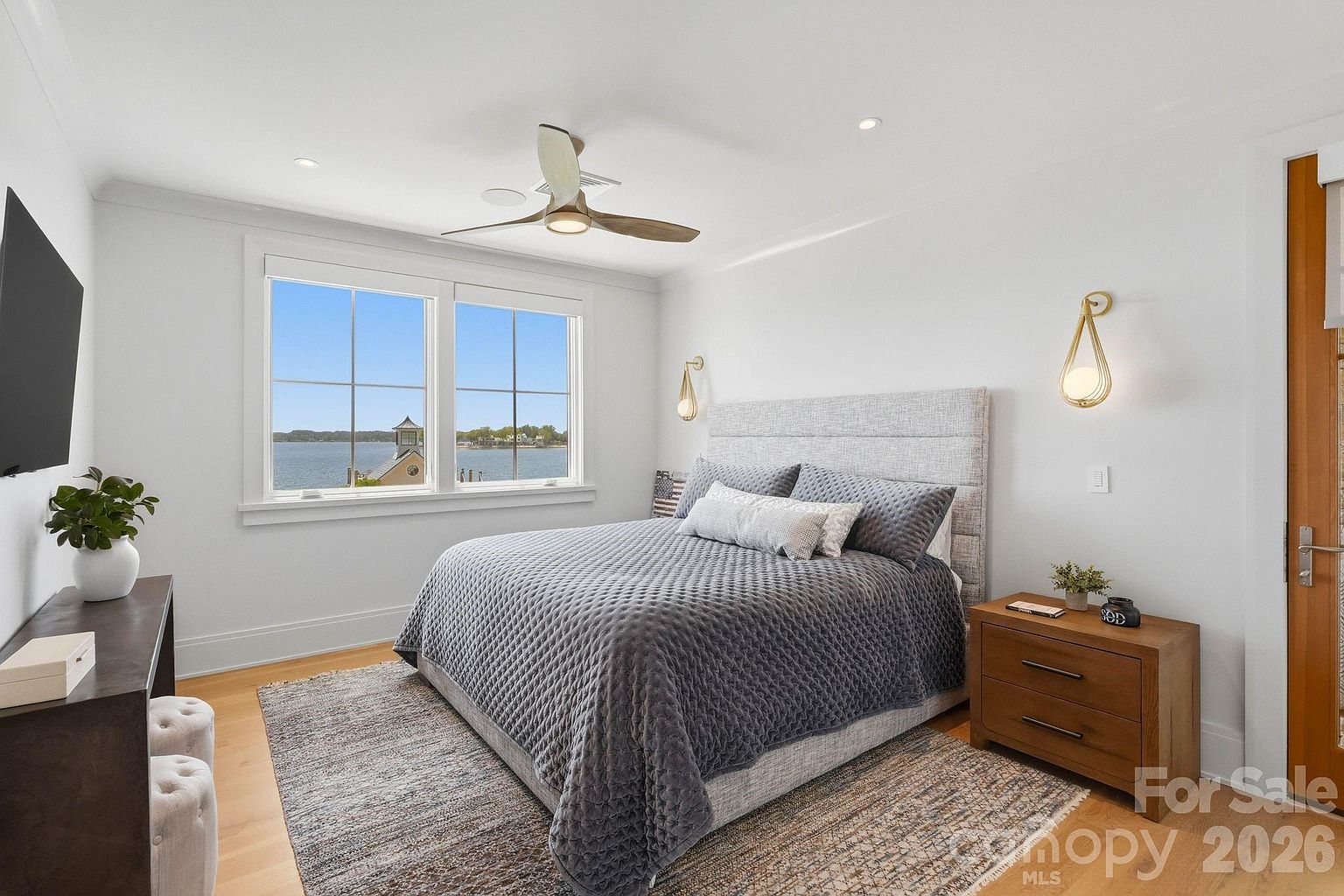 This bright and airy bedroom features a queen-sized bed with a textured gray quilt, centered against a light-colored upholstered headboard. Two elegant wall-mounted light fixtures flank the bed, while a large window provides a scenic view of the water outside. The room is completed by a wooden nightstand, a long console table with a potted plant, and a patterned area rug over light wood flooring.