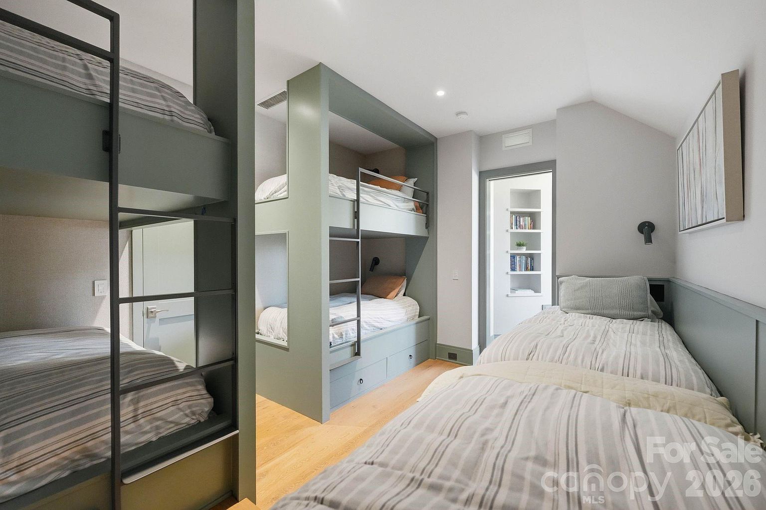 This bedroom features a space-efficient design with two sets of built-in bunk beds and a separate twin bed, all dressed in matching striped bedding. The room is finished in a soft, muted green palette with light wood flooring, creating a calm and cohesive aesthetic. A doorway in the background leads to a small alcove with built-in shelving, adding functional storage to the compact layout.