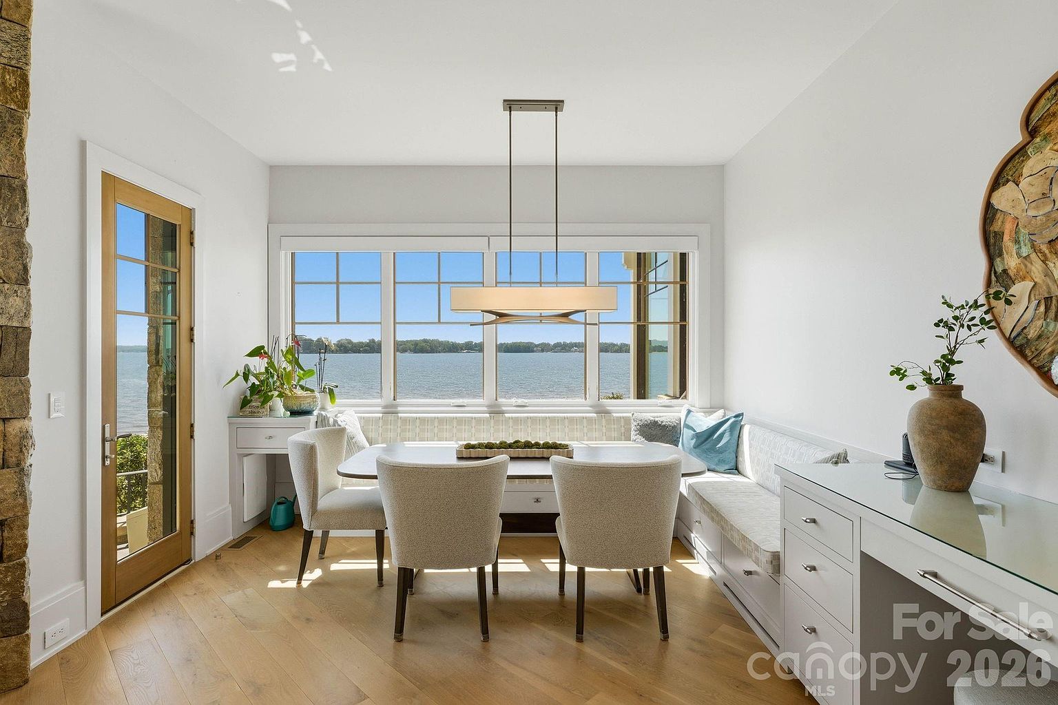 This bright dining area features a built-in banquette seating arrangement paired with a rectangular table and two upholstered chairs. Large windows provide an expansive, unobstructed view of the water, filling the space with natural light. The room is accented by a modern linear pendant light, light wood flooring, and a built-in desk or console area on the right side.