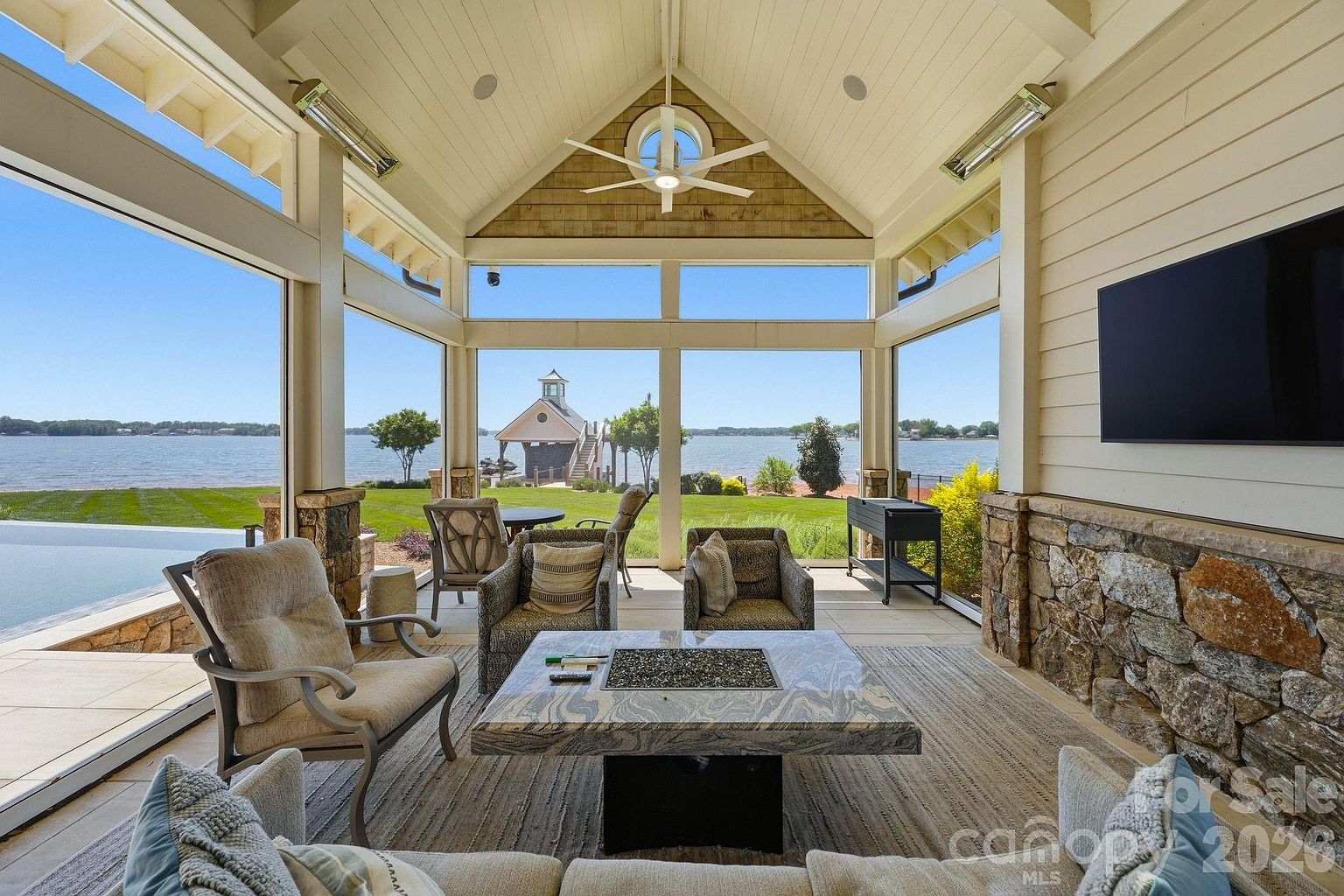 This expansive, covered outdoor living space features a vaulted ceiling with exposed beams and a central ceiling fan, creating an airy and inviting atmosphere. The area is furnished with comfortable seating arranged around a large stone-topped fire table, while a wall-mounted television and stone-clad chimney breast add functional luxury. Large glass panels provide unobstructed, panoramic views of the lake and private dock, seamlessly blending indoor comfort with the natural beauty of the waterfront.