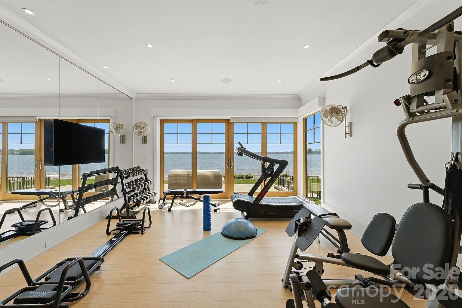 This bright and spacious home gym features light wood flooring and a wall of mirrors that reflects the room's extensive fitness equipment, including a treadmill, rowing machine, and weight station. Large windows and glass doors provide an expansive, unobstructed view of the water outside, creating an airy and motivating workout environment. The room is well-lit with recessed ceiling lights and includes a yoga mat and balance ball for versatile training.