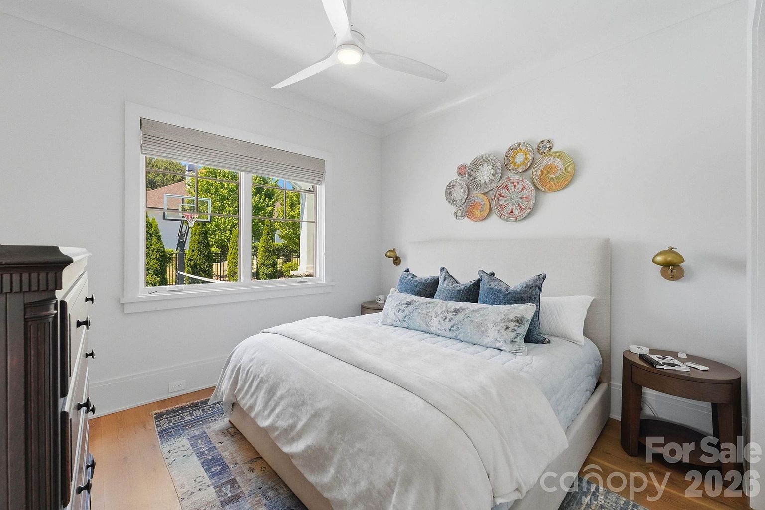 This bright and inviting bedroom features a neutral color palette with a plush upholstered headboard and layered blue-toned bedding. A decorative wall art arrangement of woven circular baskets adds texture above the bed, while a large window provides natural light and a view of the outdoor basketball court. The room is completed by a dark wood nightstand and a patterned area rug, creating a cozy and well-appointed living space.