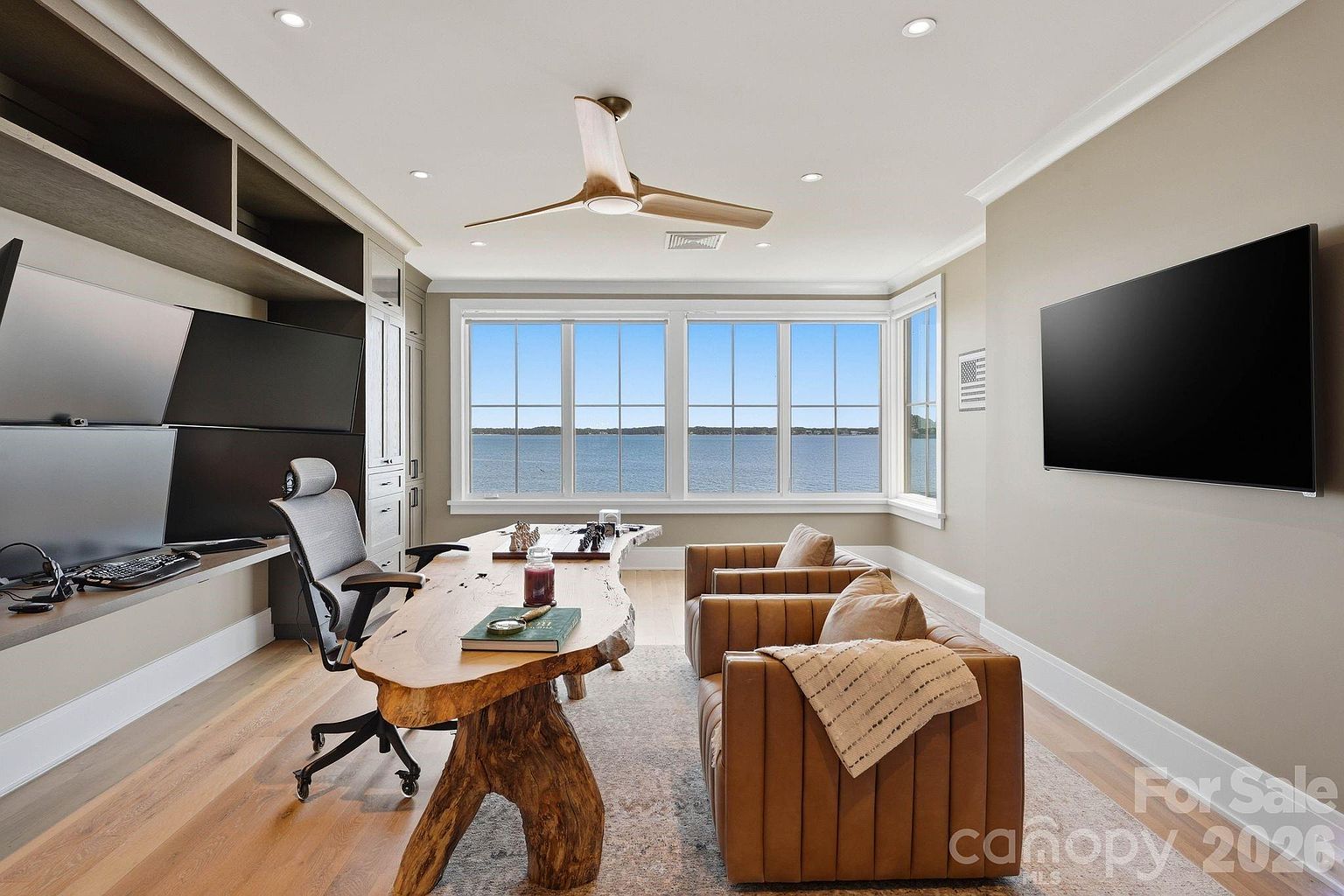 This sophisticated home office features a striking live-edge wood desk as its centerpiece, complemented by two tan leather armchairs and a modern ergonomic desk chair. Large windows provide an expansive, unobstructed view of the water, while built-in shelving and a wall-mounted television offer both functionality and entertainment. The room is finished with light wood flooring and a neutral color palette, creating a serene and professional atmosphere.