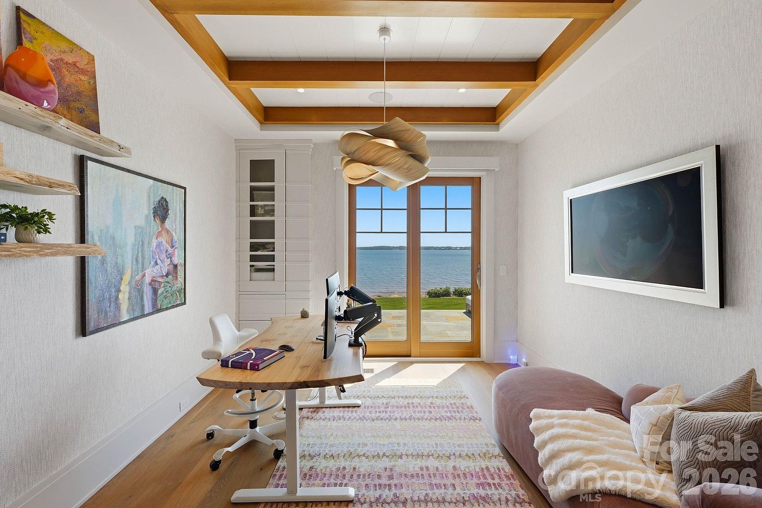This bright and modern home office features a natural wood desk, a contemporary light fixture, and a comfortable lounge area. Large glass doors provide an expansive view of the water, filling the room with natural light and creating a serene workspace. The room is accented by floating shelves, a framed piece of art, and a patterned area rug, offering a sophisticated and tranquil atmosphere.