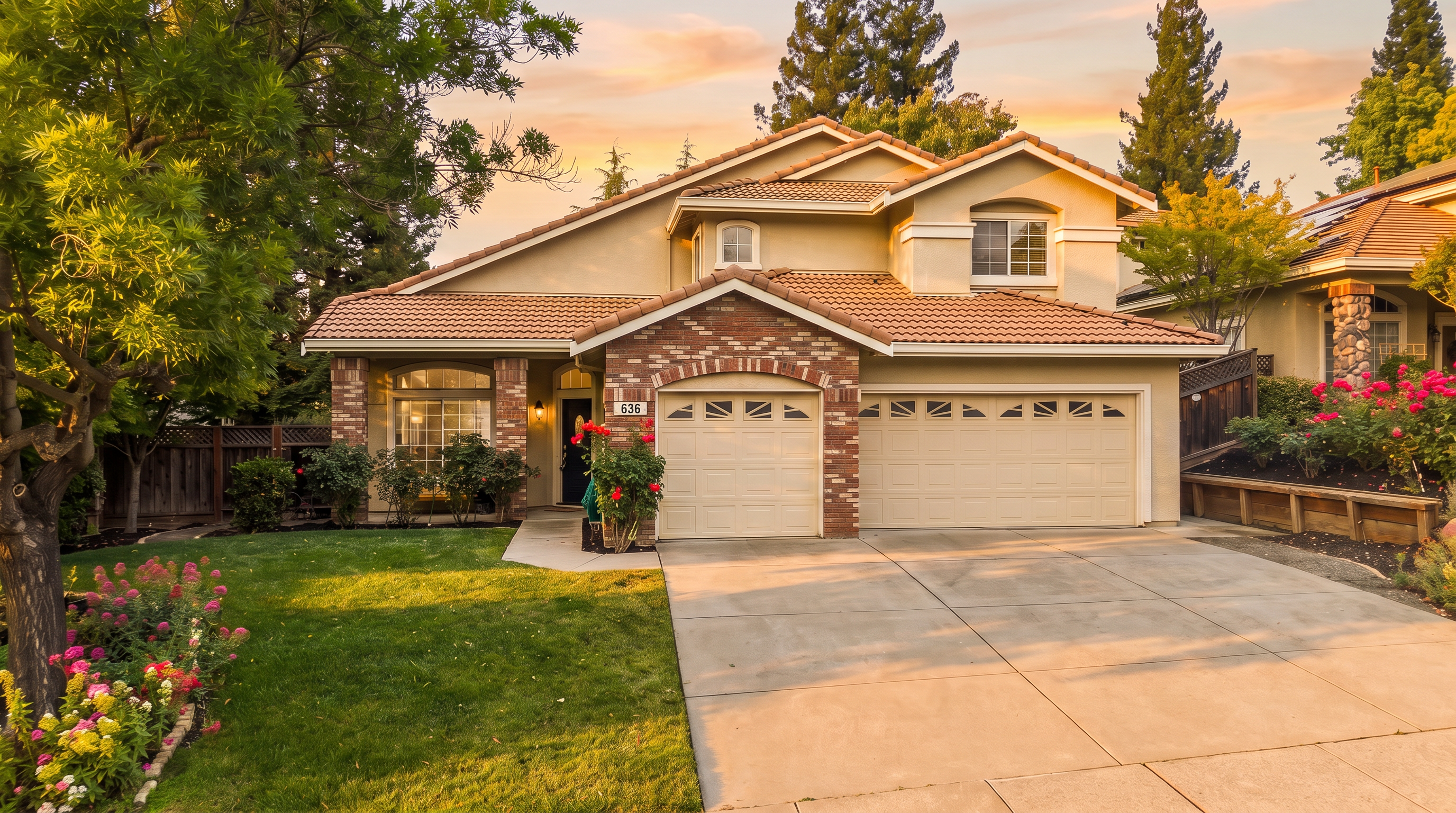 This two-story suburban home features a classic stucco facade with a tiled roof and a prominent brick-accented entryway and garage area. A well-manicured front lawn, mature trees, and vibrant rose bushes frame the property, creating an inviting curb appeal. The perspective is a slightly elevated eye-level shot, capturing the full front elevation and the concrete driveway leading to the two-car garage.