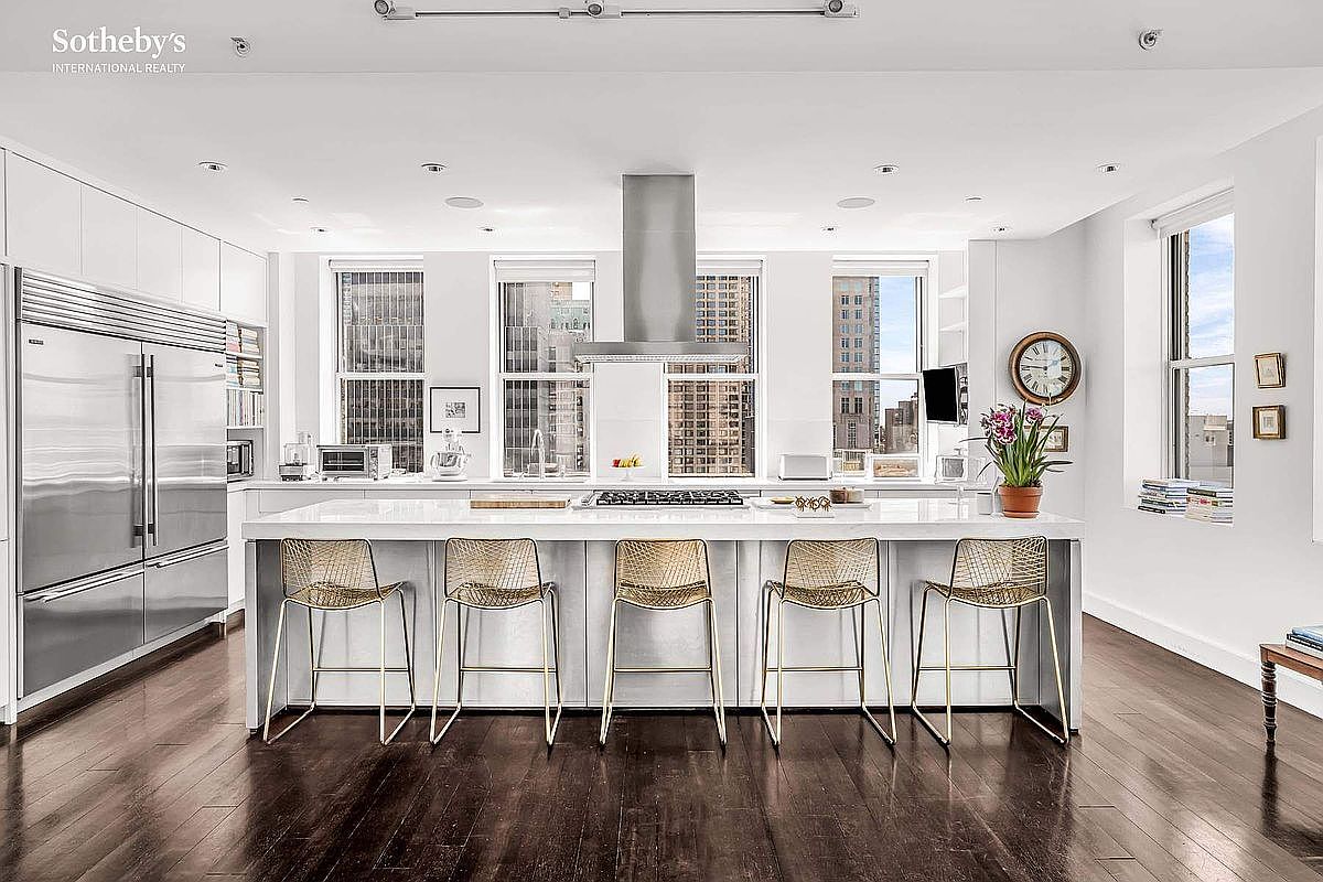 This is a bright and modern kitchen featuring a large island with seating, stainless steel appliances, and white cabinetry. The kitchen has multiple windows offering city views, and the dark hardwood floors add a touch of elegance. The overall impression is one of luxury and sophistication.