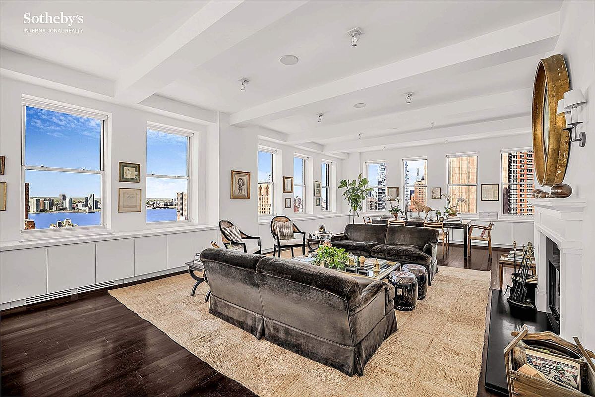 This is a spacious and elegant living room featuring hardwood floors, a neutral-toned area rug, and two plush sofas. Large windows offer stunning city and water views, while the white walls and ceiling create a bright and airy atmosphere. The room is tastefully decorated with artwork and accessories, creating a sophisticated and comfortable living space.