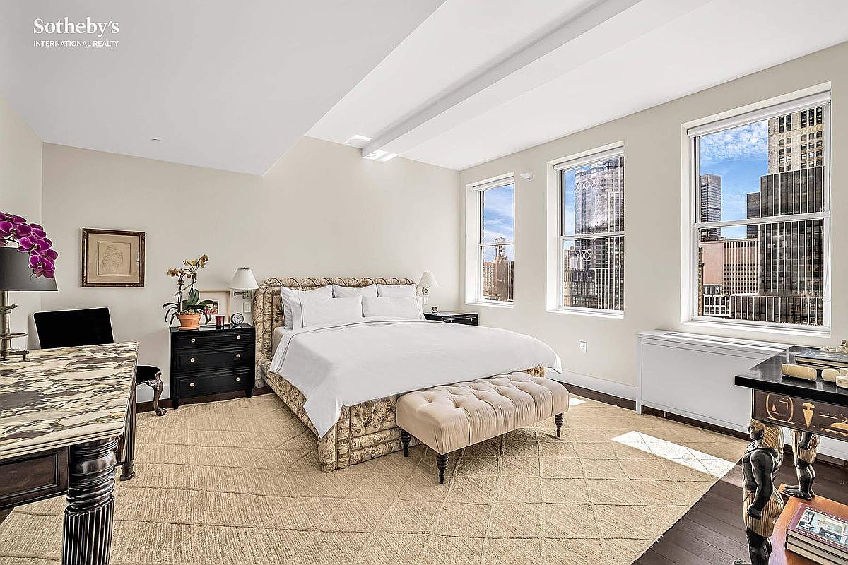 This is a primary bedroom featuring a large bed with a tufted headboard and a white duvet. The room has three windows offering city views, and there is a desk and dresser on either side of the bed. The overall style is elegant and comfortable, with a neutral color palette and a patterned rug.