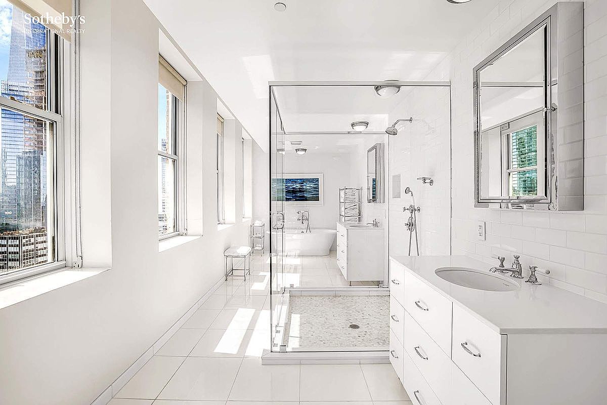 This is a bright and spacious primary bathroom featuring a glass-enclosed shower, a large vanity with ample storage, and a separate soaking tub in the background. The bathroom is predominantly white, creating a clean and luxurious feel, with large windows providing natural light and a view of the city skyline. The perspective is from the doorway, showcasing the length of the bathroom.