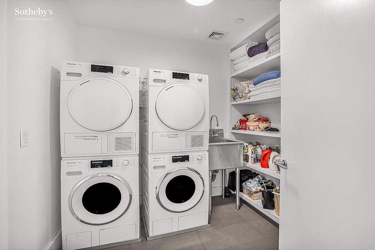 This laundry room features two stacked Miele washer and dryer units, a stainless steel utility sink with a faucet, and built-in shelving stocked with towels and cleaning supplies. The room is well-lit and appears clean and organized, offering a functional and efficient space for laundry tasks. The perspective is from the doorway, showing the full layout of the room.