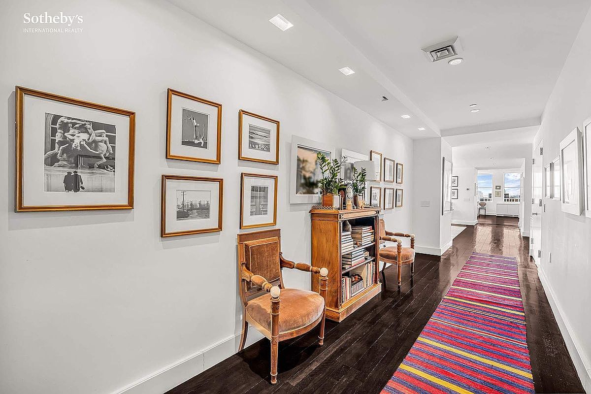 This is an interior shot of a hallway featuring dark hardwood floors and white walls adorned with framed artwork. A colorful striped runner adds a vibrant touch to the space. The hallway is furnished with a wooden bookcase and two armchairs, creating a cozy and inviting atmosphere.