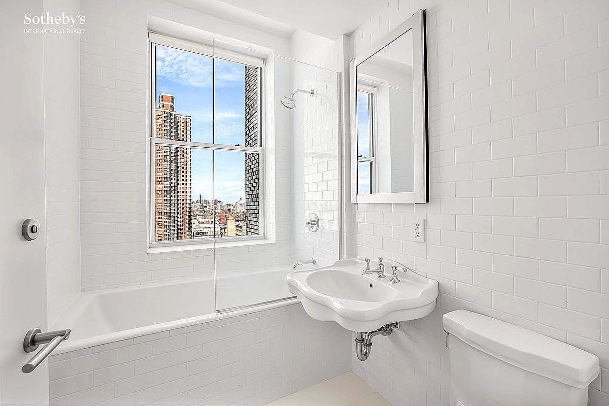 This is a bright, all-white bathroom featuring subway tile walls, a classic pedestal sink, and a large window offering a city view. The bathroom includes a bathtub with a glass partition and a modern mirror. The overall impression is clean, simple, and well-lit, emphasizing the urban view.