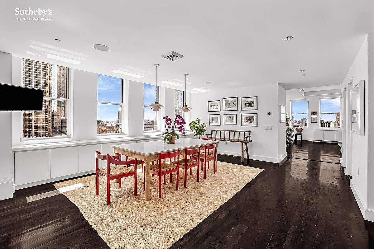 This is an interior shot of a dining room featuring a large white table with red chairs set on a circular jute rug. The room has dark hardwood floors, white walls, and large windows offering city views. Artwork adorns one wall, and pendant lights hang above the table, creating a bright and airy atmosphere.