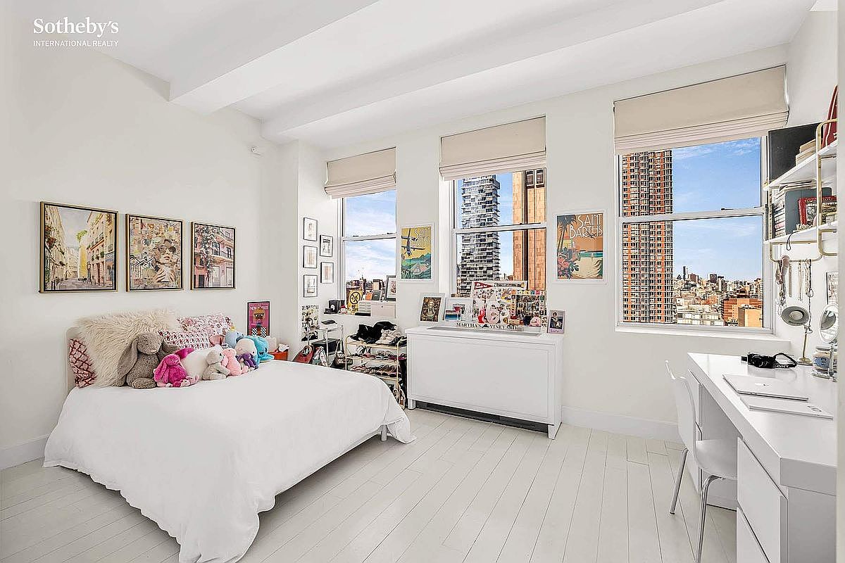 This is a bright and airy bedroom featuring white-painted hardwood floors and walls, creating a clean and spacious feel. The room is furnished with a bed adorned with plush toys and decorative pillows, complemented by artwork and personal items displayed on a console table and desk. Large windows offer city views, enhancing the room's appeal and natural light.