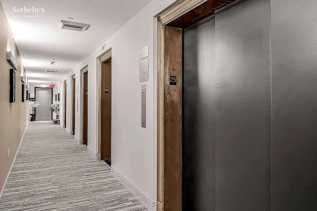 This is a well-lit hallway featuring a neutral color palette with beige walls and a patterned carpet. The hallway leads to several doors, including elevator doors marked with the number 25. The space is clean and modern, suggesting a well-maintained building.