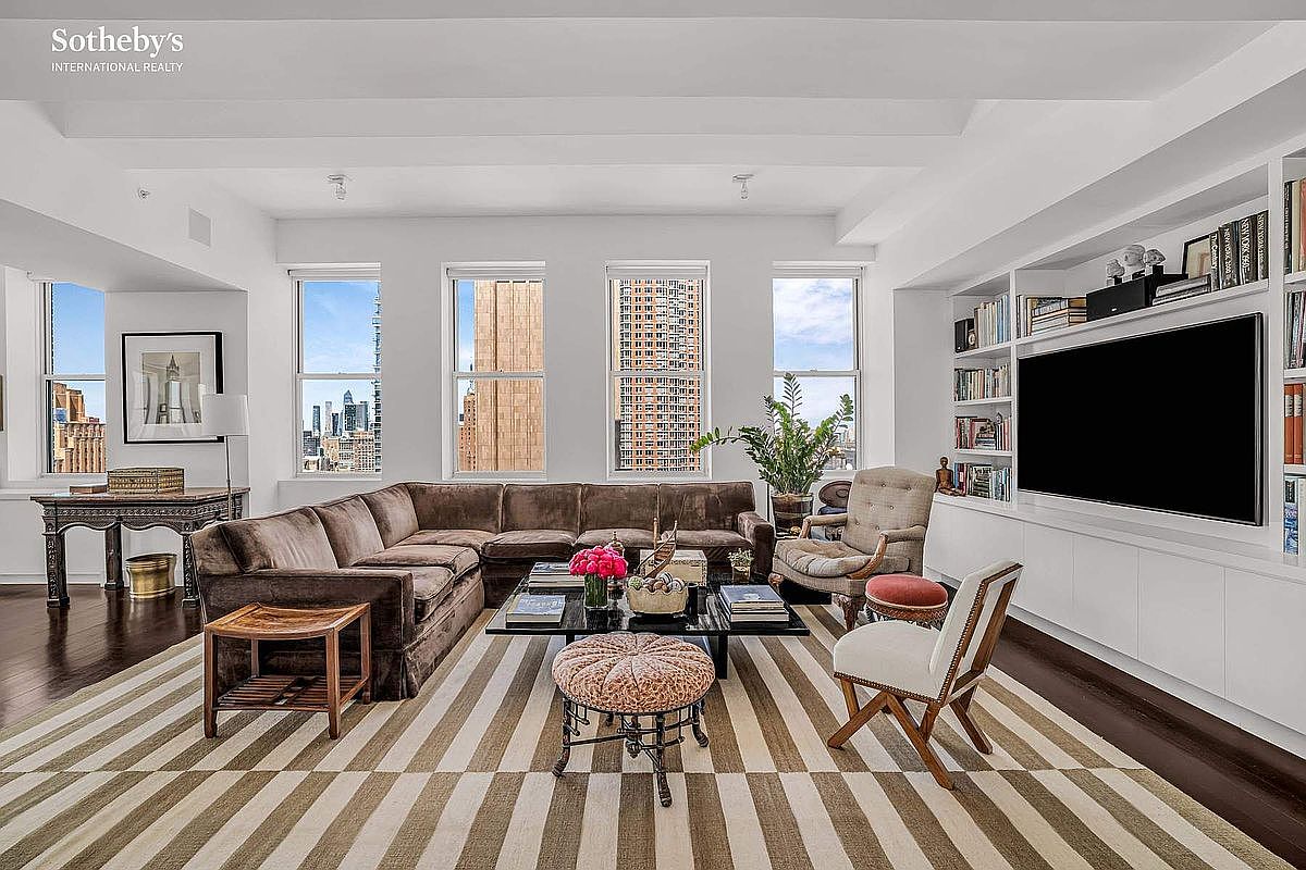 This is a spacious living room featuring a large brown sectional sofa, a glass coffee table, and a striped rug. The room has large windows offering city views and built-in shelving with a large television. The overall style is elegant and comfortable, perfect for entertaining or relaxing.