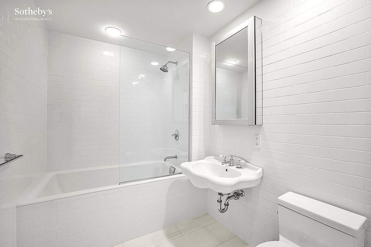 This is a bright, clean bathroom featuring white subway tiles on the walls and floor. A bathtub with a glass shower screen is visible on the left, and a wall-mounted sink and toilet are on the right. The overall impression is modern and minimalist.