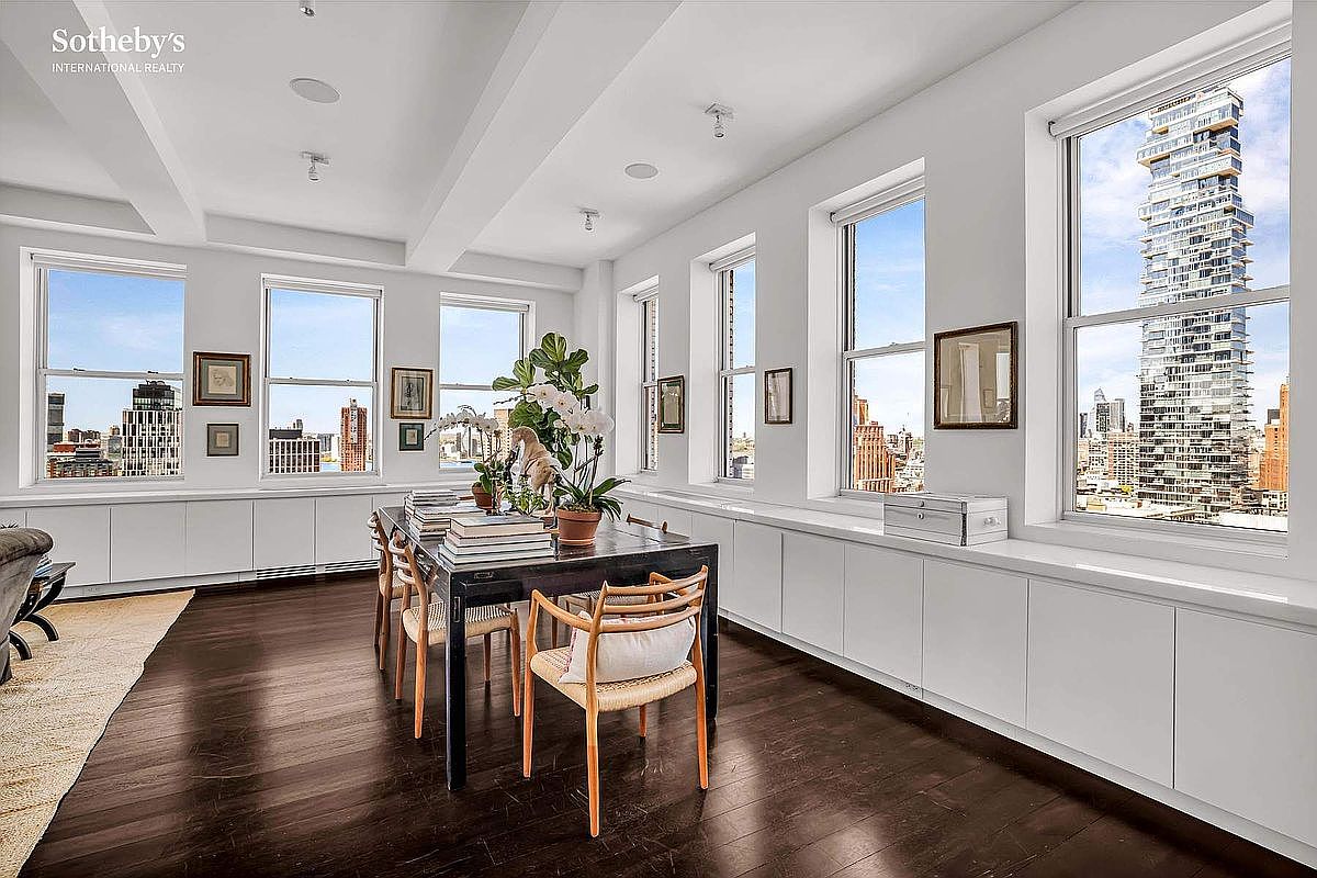 This is an interior shot of a dining room featuring a dark wood dining table surrounded by chairs with light-colored cushions. Large windows offer a stunning city view, and the room is decorated with artwork and plants. The space is bright and airy, with a modern and sophisticated aesthetic.