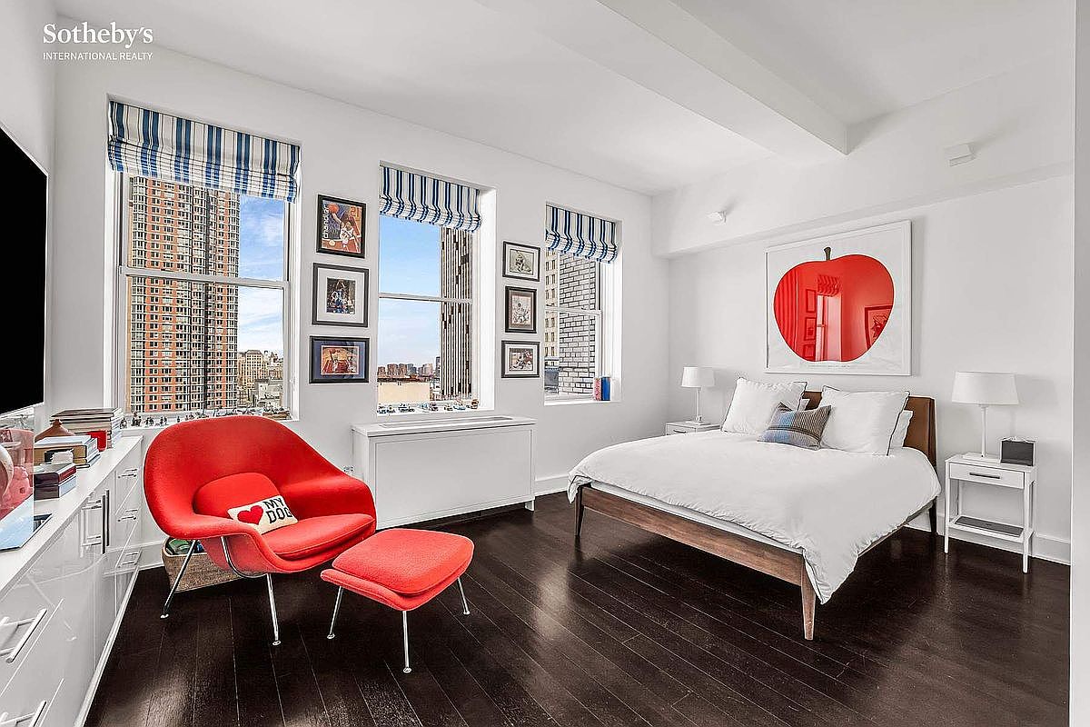 This is a bright and airy primary bedroom featuring dark hardwood floors, white walls, and large windows with city views. The room is furnished with a wooden bed dressed in white linens, a red chair and ottoman, and white bedside tables with lamps. An apple artwork hangs above the bed, adding a pop of color.