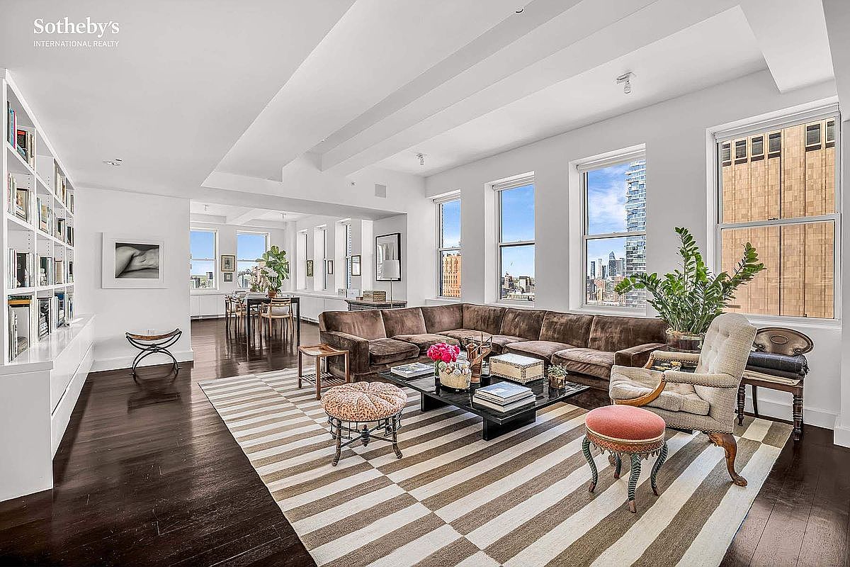 This is a spacious living room featuring a large brown sectional sofa, a striped rug, and a black coffee table. The room has multiple windows offering city views, and a bookshelf lines one wall. The overall style is elegant and comfortable, creating an inviting atmosphere.