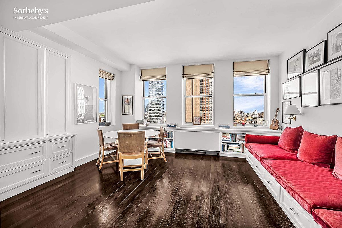This is an interior shot of a living room featuring dark hardwood floors, built-in white cabinetry and shelving, and a red cushioned seating area along the wall. Three windows offer city views, and framed artwork adorns the walls, creating a sophisticated and comfortable atmosphere. A round dining table with chairs is also present.