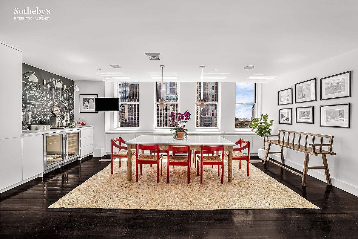 This interior shot showcases a bright and airy dining room with a modern aesthetic. The room features a large dining table with red chairs, set upon a circular patterned rug, and is illuminated by natural light streaming through large windows offering city views. A wooden bench sits against the wall adorned with framed artwork, complementing the dark hardwood flooring.
