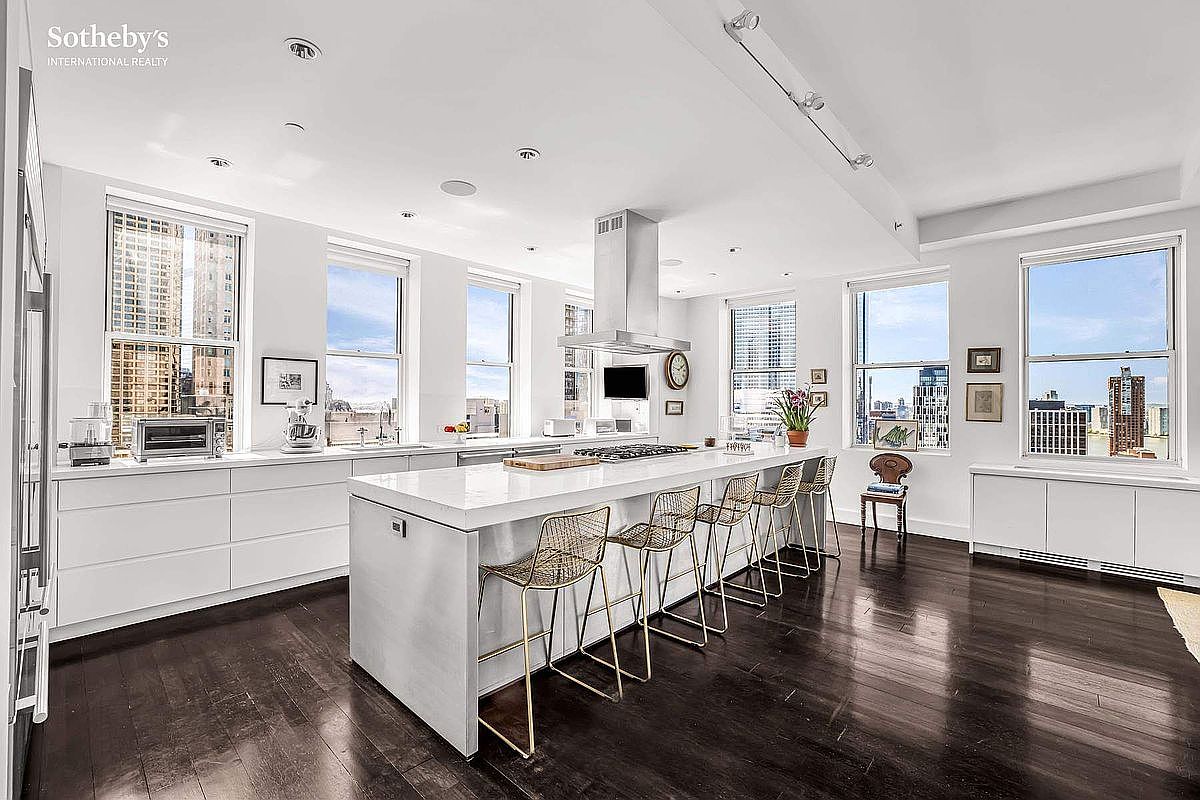 This is a spacious, modern kitchen featuring sleek white cabinetry and a large central island with seating. Stainless steel appliances and a minimalist design contribute to the contemporary aesthetic. Large windows offer city views, enhancing the bright and airy atmosphere of the space.