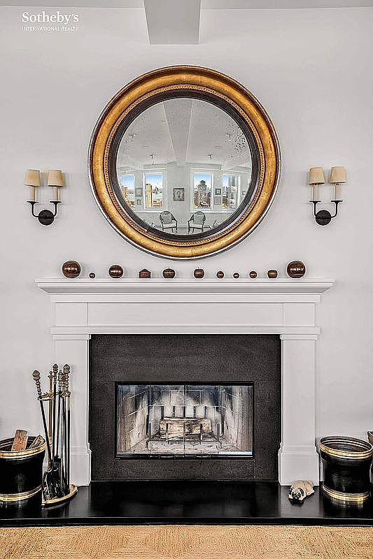 This image showcases a living room with a prominent fireplace as its focal point. The fireplace is framed by a white mantelpiece, above which hangs a large, round mirror. Flanking the mirror are sconces, and decorative spheres adorn the mantel. The overall impression is one of classic elegance and warmth.