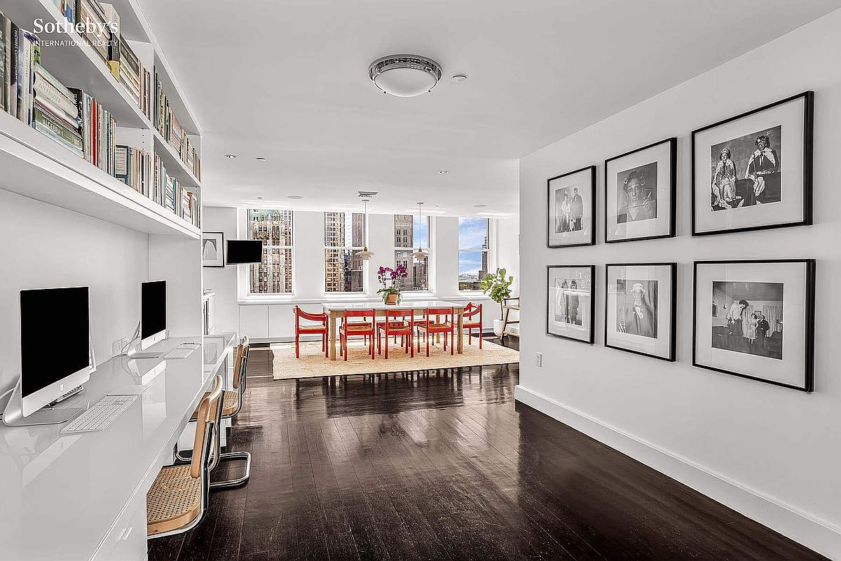This interior shot showcases a modern dining area with dark hardwood floors and white walls. A long dining table with red chairs sits on a light-colored rug, positioned between large windows offering city views. The room also features a built-in desk area with computers and a wall adorned with framed black and white photographs, creating a sophisticated and functional space.
