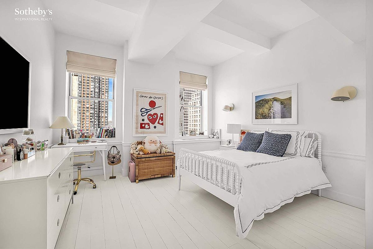 This is a bright and airy bedroom featuring white walls, white-painted hardwood floors, and two large windows that offer city views. The room is furnished with a white bed, a white dresser that doubles as a desk, and a wooden chest, creating a clean and inviting space. Artwork and decorative items add a touch of personality to the room.