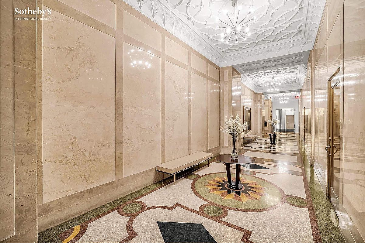 This is a luxurious hallway featuring marble walls and a decorative tile floor with a central medallion design. A bench sits against one wall, and a round table with flowers adds a touch of elegance. The ceiling is intricately designed with ornate patterns and modern lighting fixtures, creating a sophisticated and upscale atmosphere.