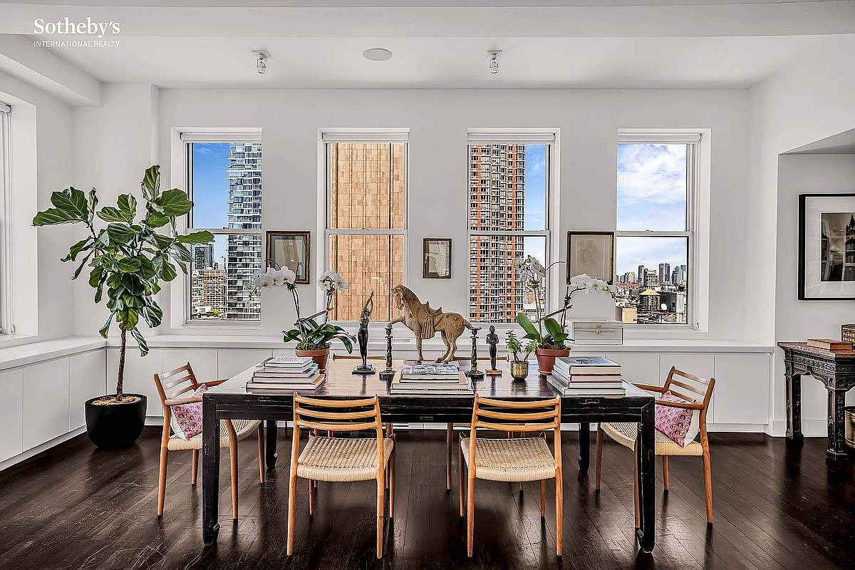 This is an interior shot of a dining room featuring a dark wood table surrounded by six chairs with woven seats. The room is bright with natural light streaming through three large windows, offering city views. Decorative items such as plants, books, and sculptures adorn the table, adding a touch of elegance and sophistication to the space.