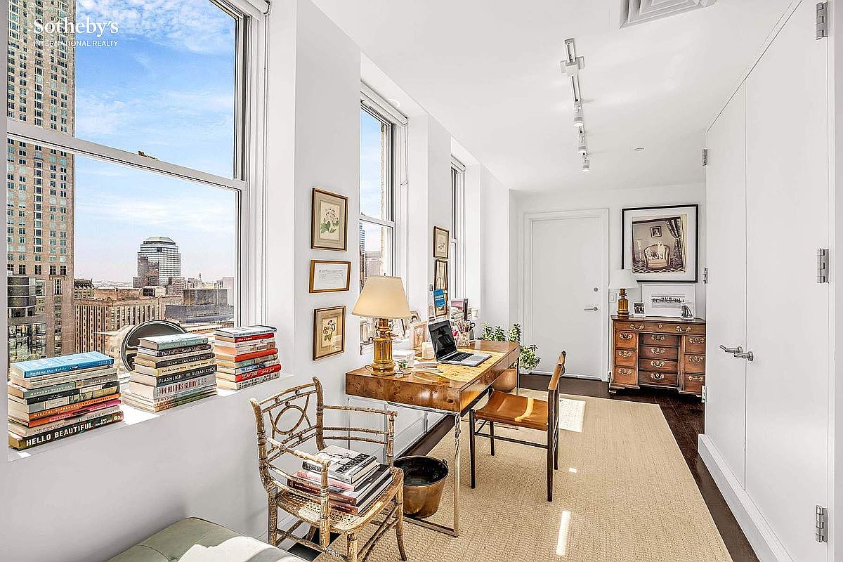 This is an interior shot of a bright and airy office space. The room features large windows offering city views, a wooden desk with a laptop, and a mix of modern and vintage furniture. Books are stacked on the windowsill and a woven rug covers the floor, creating a cozy and sophisticated atmosphere.
