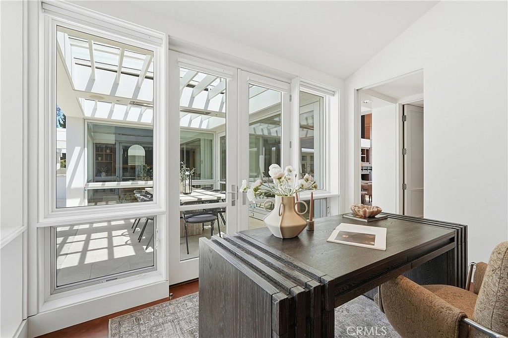 This bright home office features a dark wood desk positioned to overlook an adjacent sun-drenched courtyard through large glass doors and windows. The room is decorated with a minimalist aesthetic, highlighted by a decorative vase of flowers on the desk and a patterned area rug. The perspective looks from the office interior toward the outdoor space, emphasizing the seamless connection between indoor and outdoor living areas.