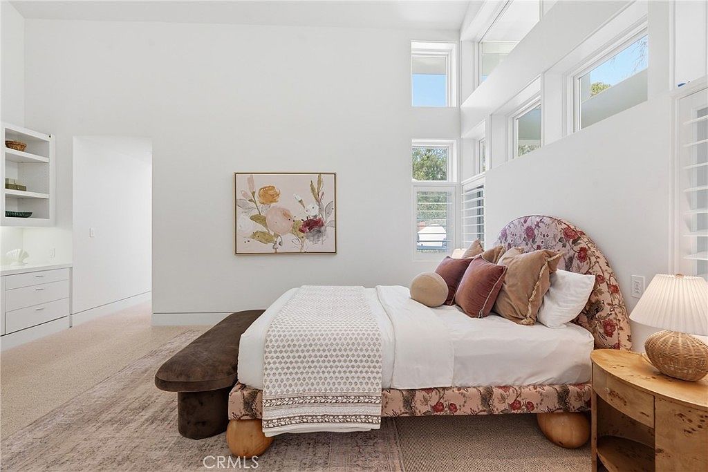 This bright and airy primary bedroom features a high-ceiling design with large windows that allow for abundant natural light. The room is anchored by a bed with a distinctive floral-patterned headboard and frame, complemented by a textured bench at the foot and a light wood nightstand. A piece of floral artwork hangs on the wall, adding a soft, decorative touch to the minimalist white interior.