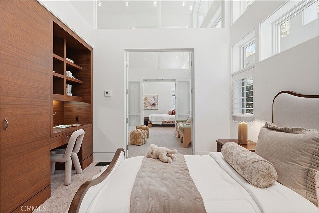 This bright and airy bedroom features a neutral color palette with a built-in wooden desk unit and shelving integrated into the wall. A plush bed with a soft-toned headboard sits in the foreground, while an open doorway leads to an adjacent room, creating a sense of depth and connectivity. The room is illuminated by natural light from tall windows and features high ceilings, contributing to an open and sophisticated atmosphere.