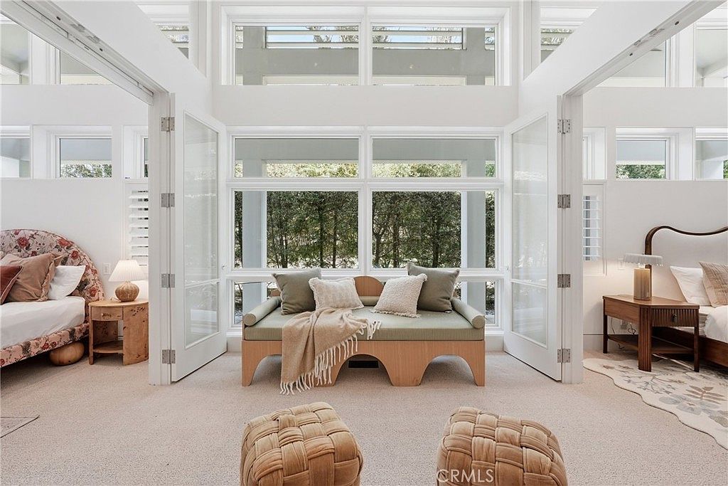 This bright and airy bedroom features a symmetrical layout with two beds flanking a central seating area. Large windows with white frames span the rear wall, allowing ample natural light to fill the space, while woven ottomans and a low-profile bench provide comfortable seating. The room is decorated in a neutral, soft palette, creating a serene and inviting atmosphere.