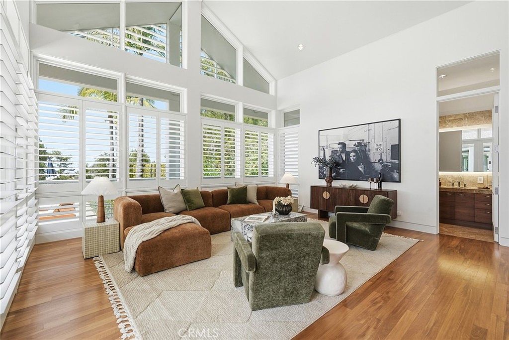 This spacious living room features a high, vaulted ceiling and a wall of large windows with white plantation shutters that flood the space with natural light. The room is anchored by a large, brown sectional sofa and two olive-green armchairs arranged on a textured area rug over warm-toned wood flooring. A dark wood console table sits against the back wall, topped with a large framed black-and-white photograph, creating a sophisticated and airy atmosphere.