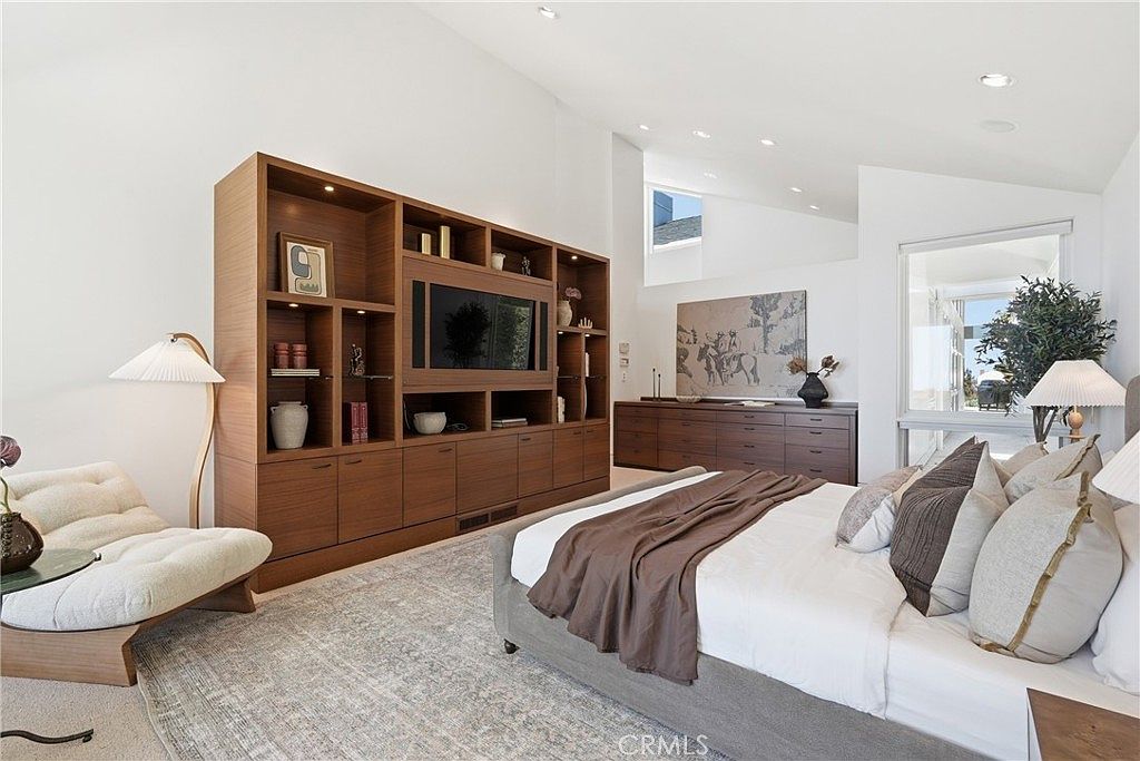 This spacious primary bedroom features a large, light-toned bed with a brown throw blanket, positioned in front of a custom wood media unit with integrated shelving. To the left, a modern lounge chair sits beside a tall, minimalist floor lamp, while a long dresser with a large piece of artwork sits against the opposite wall. The room is brightened by large windows that provide a view of the outdoors and a high, vaulted ceiling that enhances the airy, contemporary atmosphere.