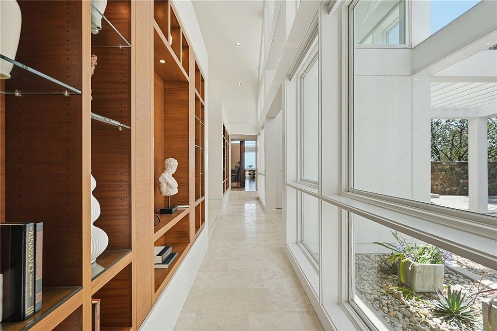 This bright, modern hallway features a long wall of custom light wood built-in shelving and cabinetry, accented by decorative sculptures and books. To the right, large windows provide an expansive view of an outdoor garden area with stone landscaping and greenery. The polished stone flooring reflects the natural light, creating an airy and sophisticated transition space within the home.