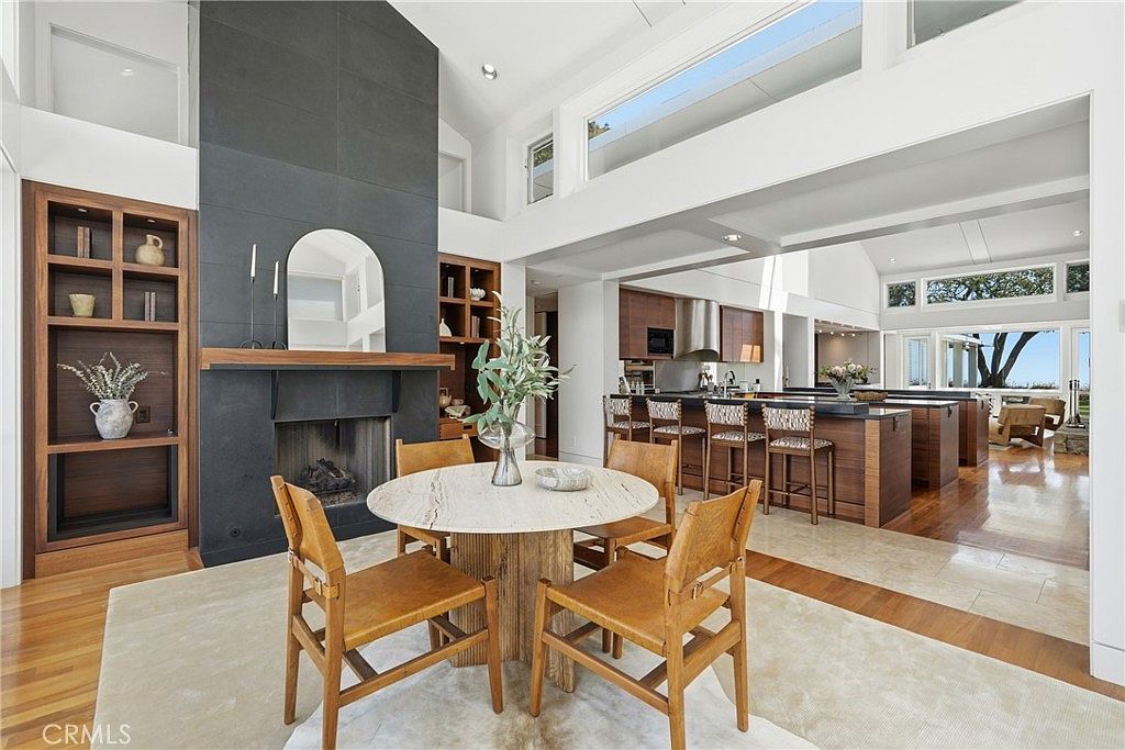 This open-concept dining area features a round stone-topped table surrounded by four leather-upholstered chairs, set atop a neutral area rug. A prominent dark-toned, full-height chimney breast serves as a focal point, flanked by built-in wooden shelving units. The space flows seamlessly into a bright kitchen with a large island and bar seating, all illuminated by natural light from high-placed windows.