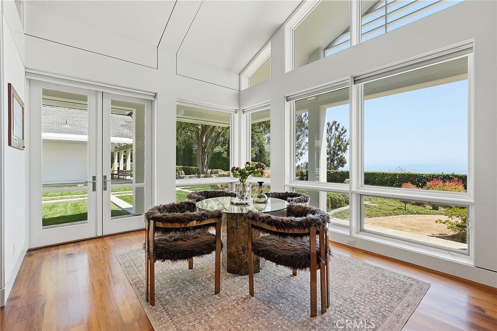This bright dining area features a glass-topped table surrounded by four chairs upholstered in textured, dark faux-fur fabric. The room is defined by expansive windows that offer panoramic views of the surrounding landscape and sky, creating a seamless connection to the outdoors. A patterned area rug anchors the space, which is finished with warm-toned wood flooring and crisp white walls.