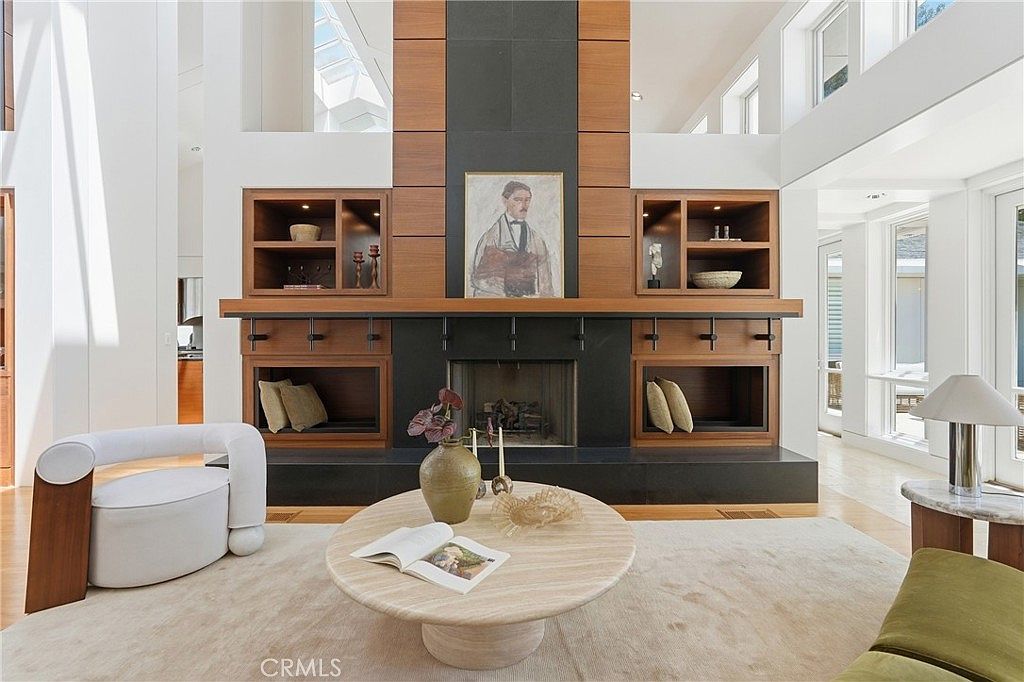 This spacious living room features a prominent central fireplace with a dark, full-height surround flanked by built-in wood shelving units. A circular stone coffee table sits in the center of the room on a light-colored area rug, complemented by a modern white armchair and a green sofa. The room is characterized by its high, vaulted ceilings and large windows that allow for abundant natural light, creating an airy and sophisticated atmosphere.