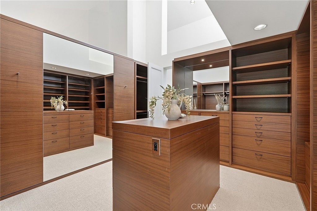 This spacious walk-in closet features extensive custom wood cabinetry with integrated shelving and drawers, providing ample storage. A central island with a built-in electrical outlet serves as a focal point, while a large mirror reflects the room to enhance the sense of scale. The room is finished with neutral carpeting and tall, clean lines, creating a sophisticated and organized atmosphere.